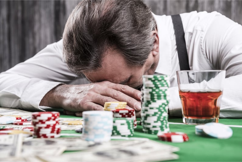 Depressed senior man in shirt and suspenders leaning his head at the poker table with money and gambling chips laying all around him